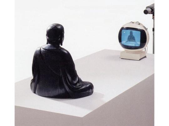 a statue of a Buddha views its own image on a television